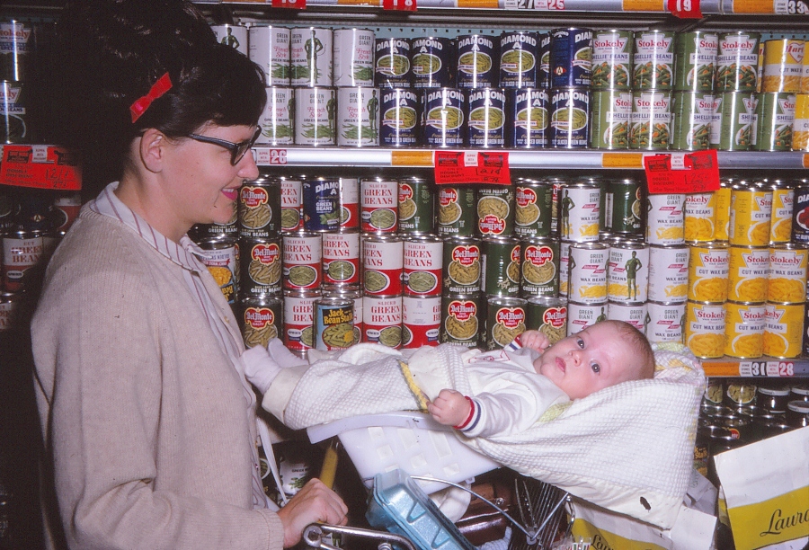 A mother (Silent Generation) pushes a Gen-X baby in a shopping cart. The baby is straddling the flip-up child seat. 