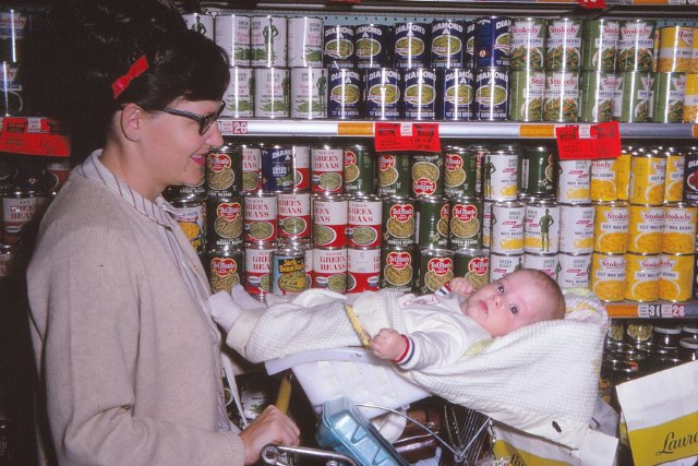 A mother (Silent Generation) pushes a Gen-X baby in a shopping cart. The baby is straddling the flip-up child seat.